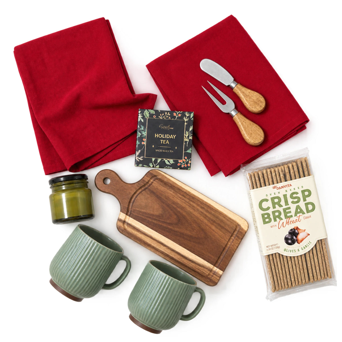 Flat lay view of I'll Be Home Christmas gift box contents featuring two sage green ribbed ceramic mugs, solid acacia wood charcuterie board with cheese knives, crispbread, spiced holiday tea, red kitchen towels, and green jar candle on white background