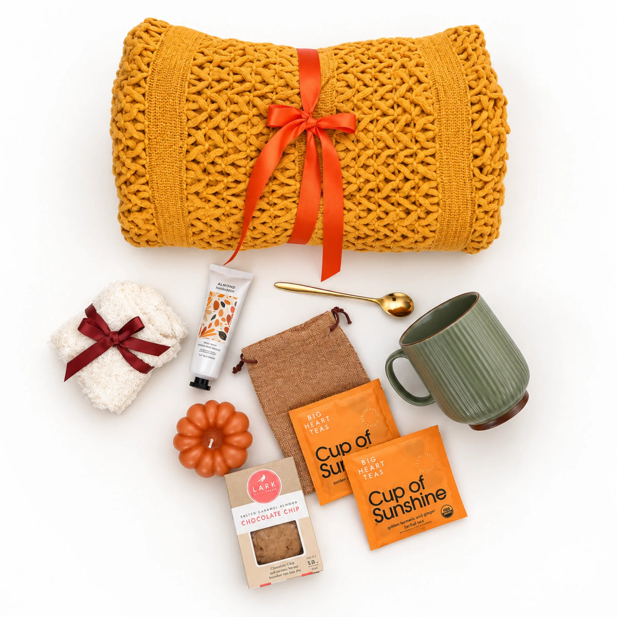 Flat lay Pumpkin Glow gift box items showing chunky throw blanket, sage mug, pumpkin candle, hand cream, tea sachets in linen pouch, socks, and gold spoon on white background