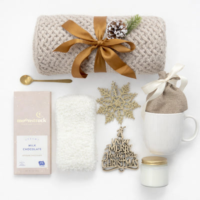 All I Want is You Gift Box flat lay featuring beige knitted throw blanket with gold ribbon, white ceramic mug, glass candle with gold lid, golden coffee spoon, Moonstruck milk chocolate, white fluffy socks, burlap gift bag, and gold Christmas ornaments on pure white background.