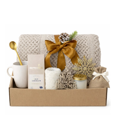 All I Want is You Gift Box holiday set arranged inside open kraft box with beige knitted throw blanket, white ceramic mug, Moonstruck milk chocolate, white fluffy socks, glass candle with gold lid, burlap bag, golden spoon, and festive Christmas ornaments.