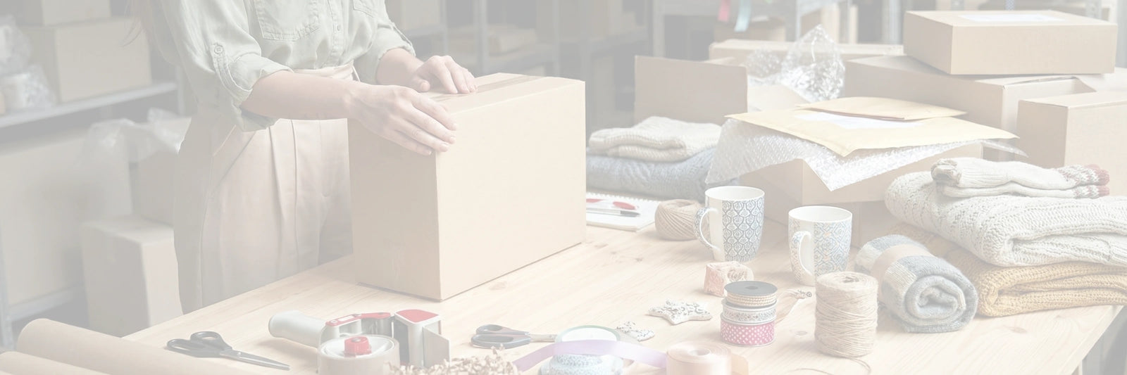 A behind-the-scenes look at the Arinna Gifts Dallas warehouse, showing hands carefully taping shut a shipping box on a table filled with curated items like mugs and cozy knits, ready for delivery.