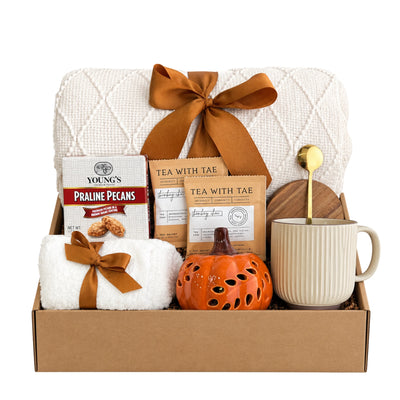 Autumn Is Calling gift box featuring cream diamond knit blanket, ceramic pumpkin luminary, ribbed ceramic mug, acacia wood coaster, praline pecans, chai tea sachets, and plush fuzzy socks
