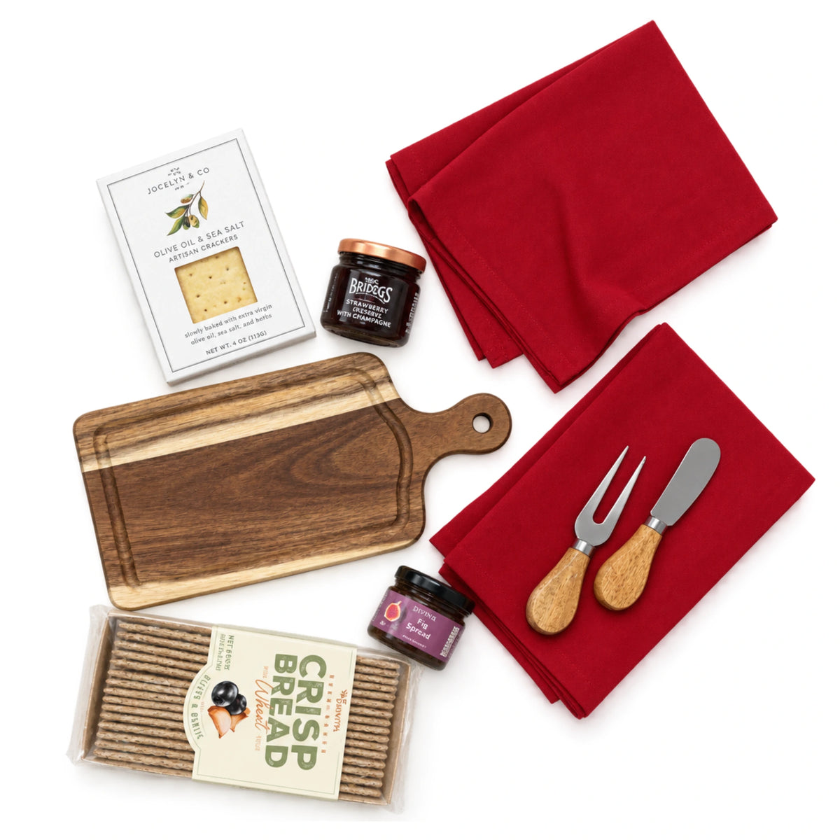 Flat lay view of Christmas Eve gift box items showing acacia cutting board, cheese fork and knife, olive oil crackers, crispbread, fig spread, jam, and red napkins on white background