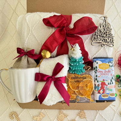 Christmas Eve Gift Box by Arinna Gifts featuring a throw blanket, festive socks, gourmet chocolate, coffee mug, elegant spoon, hot chocolate, candle, and Christmas ornaments.