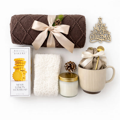 Flat lay of Christmas Morning Gift Box items including brown knit blanket, holiday ornament, Meyer Lemon cookies, candle, ceramic mug, fluffy socks and hot chocolate pouch.