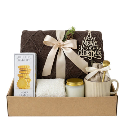 Christmas Morning Gift Box featuring brown knit throw blanket, Meyer Lemon cookies, glass jar candle, fluffy socks, ceramic mug with gold spoon and hot chocolate in burlap bag.