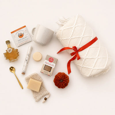 Flat lay Hello Fall gift box with cozy knit blanket, ceramic mug, maple syrup bottle, cookies, herbal tea, soap saver pouch and gold spoon arranged on white background