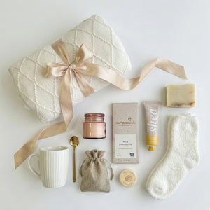 Cream cozy gift box items flatlay featuring a knitted blanket with satin ribbon, white mug, gold spoon, shea hand cream, artisan soap, pink candle, chocolate bar, fluffy socks, and burlap pouch displayed on a clean white background.