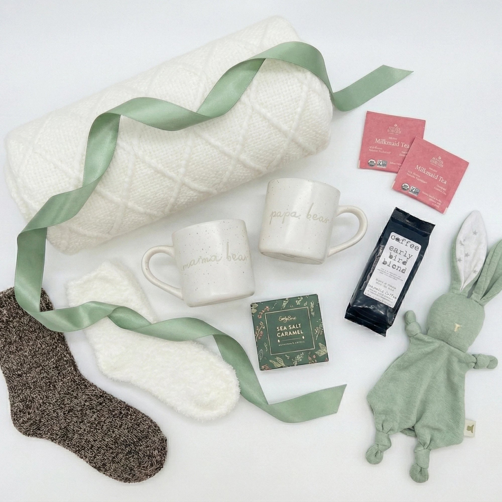 Parents Relax Box flatlay featuring a white knitted blanket, mama and papa bear mugs, cozy socks, herbal tea packets, artisan coffee, sea salt caramel chocolates, and a soft green bunny lovey arranged on a clean white background