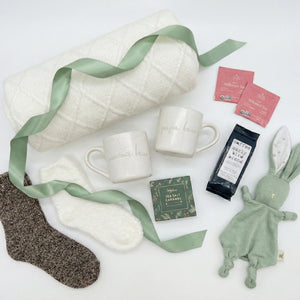 Parents Relax Box flatlay featuring a white knitted blanket, mama and papa bear mugs, cozy socks, herbal tea packets, artisan coffee, sea salt caramel chocolates, and a soft green bunny lovey arranged on a clean white background