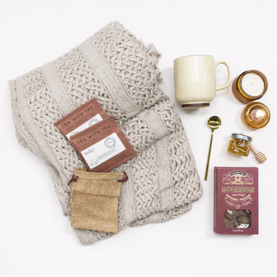 Flat lay of Quiet Comfort Gift Box care package for women featuring cozy blanket, herbal tea, honey, candle and ceramic mug